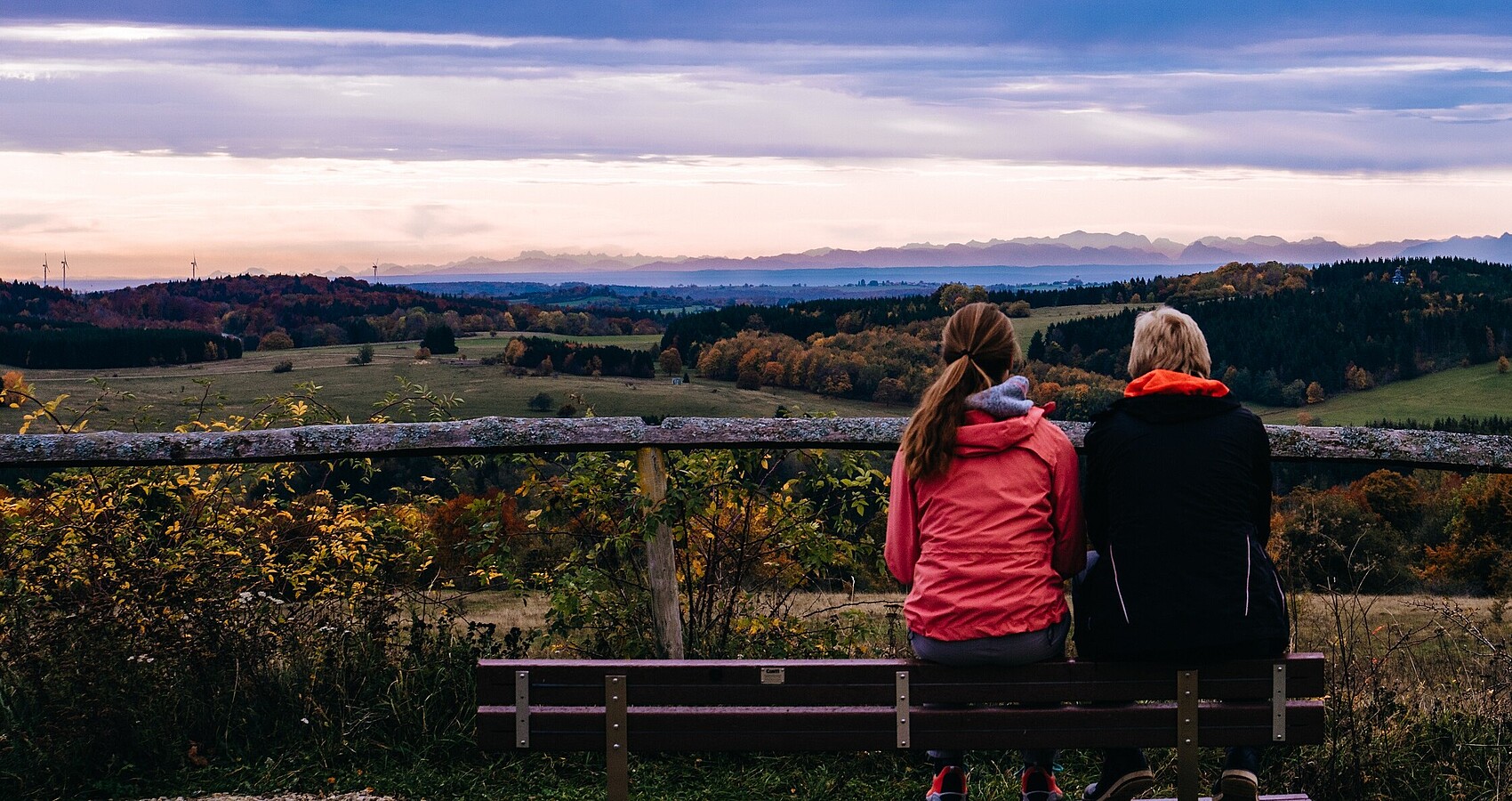 Zwei Frauen sitzen bei Sonnenaufgang auf einem Berg auf einer Bank und schauen ins Tal