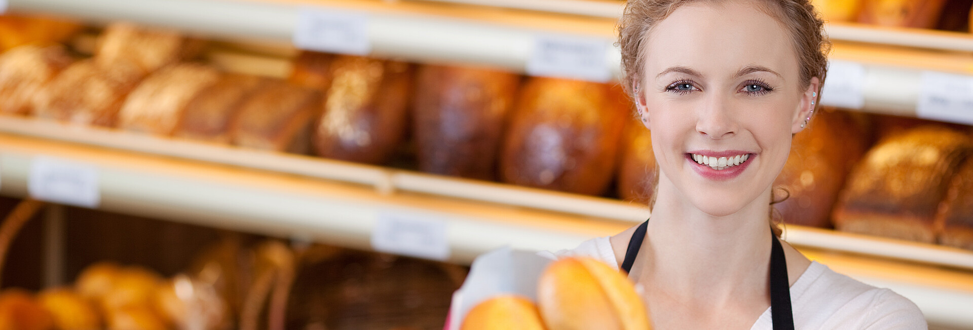 Eine Bäckereiverkäuferin hält dem Betrachter lächelnd eine Tüte mit Brötchen entgegen