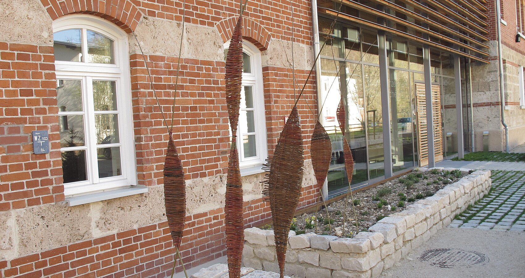 Das Foto zeigt selbstgemachten Deko-Stelen aus Weide in Rabatten vor einem Backsteinhaus