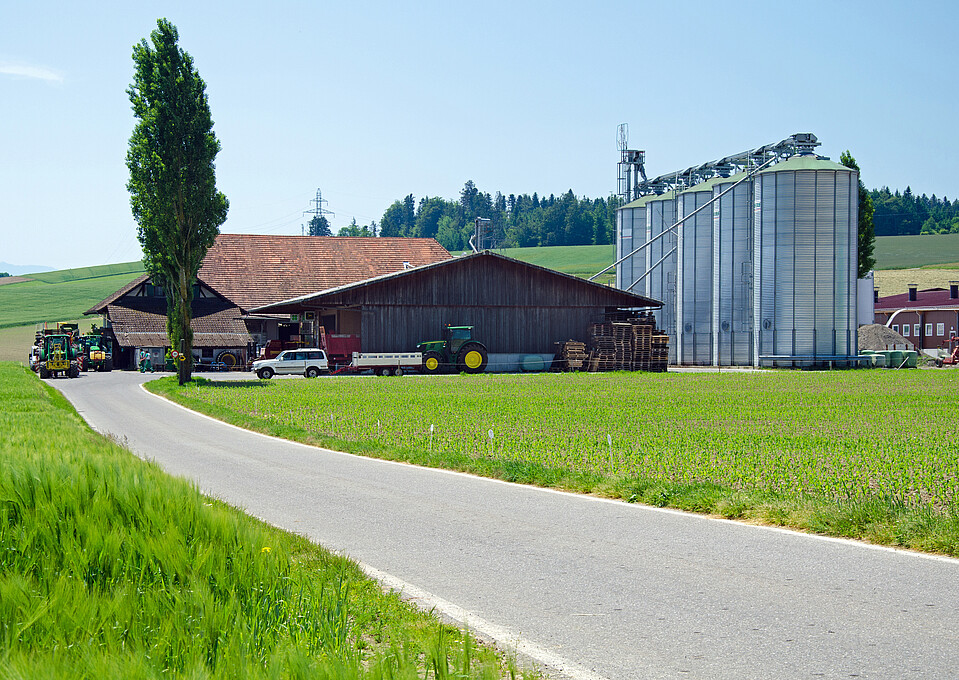 Man sieht einen Bauernhof mit Betriebsgebäuden, Maschinen und umgeben von grünen Ackerbauflächen