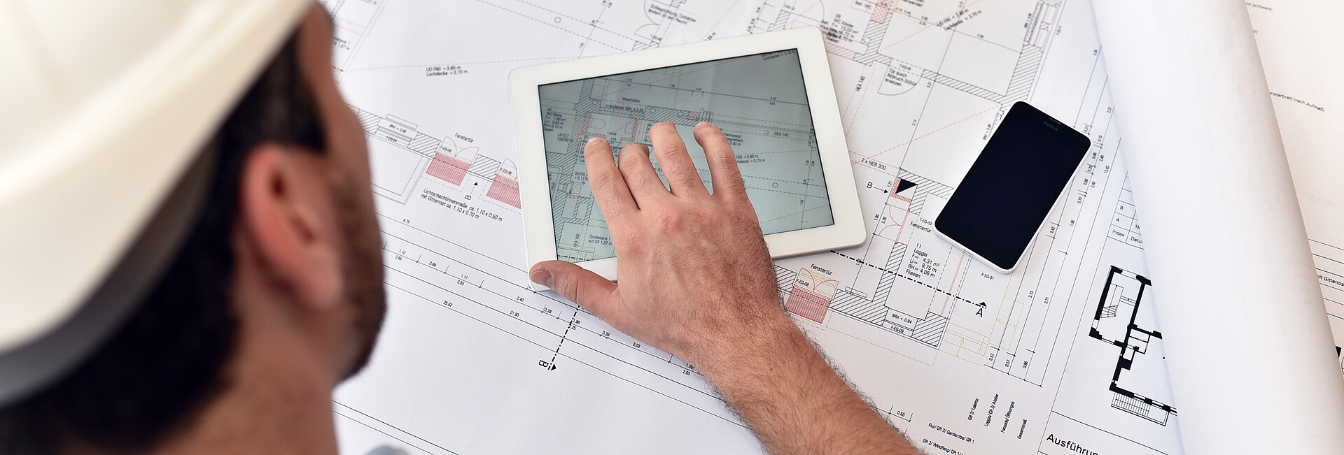 Ein Bauzeichner sitzt mit Tablet, handy und Zeichenutensilien an einem Plan